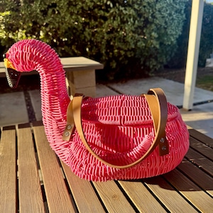 May include: A pink woven flamingo-shaped handbag with brown leather handles. The bag is made of woven material and has a detailed design. The flamingo's head is upright, with a yellow beak and black accents. The bag sits on a wooden table.