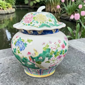 May include: A white ceramic ginger jar with a matching lid, featuring a colorful hand-painted floral and bird motif. The design includes pink, yellow, and blue flowers, green foliage, and a bird, with a blue decorative band and a yellow base rim.