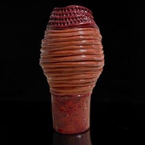 May include: A ceramic vase with a cylindrical base and a bulbous body. The base is a textured dark red, while the upper body features horizontal clay coils in shades of brown and orange. The rim has a dark red, honeycomb-like textured pattern.
