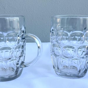 Op de afbeelding: Twee heldere glazen bierpullen met een textuur, kuiltjesontwerp. Elke pul heeft een handvat en een 0,5L markering. De pullen zijn leeg en staan op een witte ondergrond. De achtergrond is lichtgrijs.