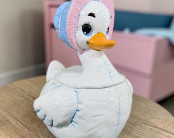 May include: A white ceramic duck figurine with a glossy finish, featuring a blue and pink bonnet, an orange beak, and large black eyes. The duck has molded details resembling fabric seams and is shaped as a lidded container with a rounded body.