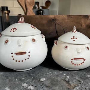 May include: Two white ceramic jars with lids, each adorned with a face design. The faces feature brown eyes, a mouth, and dotted details. The lids have a decorative finial and floral accents. These jars are ideal for kitchen storage.