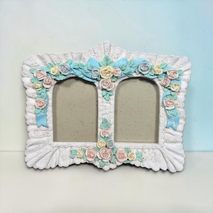 May include: A white double photo frame with a scalloped edge and floral decorations. The frame has two arched openings, decorated with pastel-coloured roses, ribbons, and bows in shades of pink, yellow, and blue. The frame is made of a solid material.