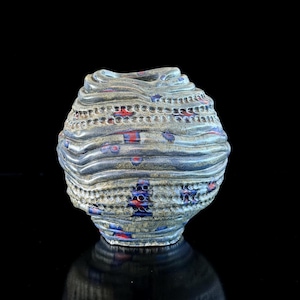 Può includere: Un vaso in ceramica fatto a mano con un design unico e strutturato. Il vaso presenta creste orizzontali e sezioni perforate, con accenti rossi e blu. Il colore generale è un grigio-blu tenue, che crea un'estetica moderna e artistica.