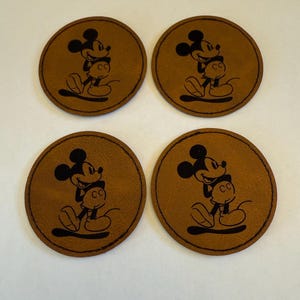Könnte beinhalten: Vier braune Lederuntersetzer mit einem schwarzen Mickey Mouse-Design. Jeder Untersetzer hat eine runde Form mit einem genähten Rand und einer detaillierten Illustration von Mickey Mouse.