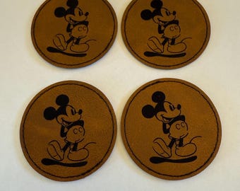 Op de afbeelding: Vier bruine leren onderzetters met een zwart Mickey Mouse-ontwerp. Elke onderzetter heeft een ronde vorm met een gestikte rand en een gedetailleerde illustratie van Mickey Mouse.