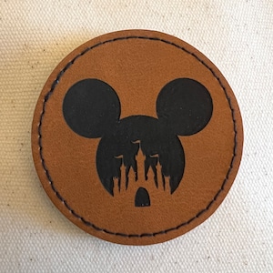 May include: A brown leather coaster with a black silhouette of Mickey Mouse's head with the silhouette of Cinderella's castle inside the ears.