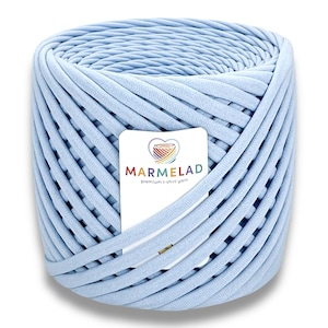 Puede incluir: Una gran madeja cilíndrica de hilo de camiseta azul claro. El hilo está bien enrollado, con bucles visibles y una textura suave. Una etiqueta blanca con el texto "MARMELAD premium t-shirt yarn" está adjunta.