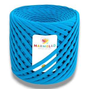 Peut inclure: Une bobine cylindrique de fil de t-shirt turquoise vif. Une étiquette blanche avec le texte "MARMELAD premium t-shirt yarn" est enroulée autour du fil. L'étiquette présente un logo coloré avec un motif arc-en-ciel.