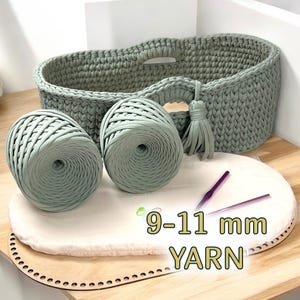 May include: A sage green crochet basket with two matching yarn balls, a wooden base, and crochet hooks. The text "9-11 mm YARN" is visible. The basket has handles and a tassel. The yarn is suitable for 9-11 mm crochet projects.