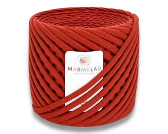 May include: A large, cylindrical spool of rust-colored t-shirt yarn. A white label with the word "MARMELAD" in orange letters is wrapped around the yarn. The label also features a heart-shaped logo with a rainbow design.