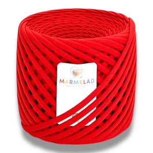 Peut inclure: Une pelote de laine rouge vif, enroulée en forme cylindrique. Une étiquette blanche avec le mot "MARMELAD" et un logo est attachée. La laine est un matériau épais et doux, idéal pour le tricot, le crochet et d'autres projets créatifs.