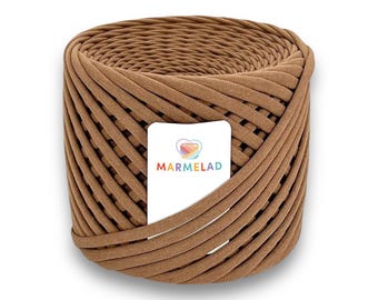 May include: A tightly wound roll of brown fabric yarn. The yarn is a light brown color and appears to be made of a soft, textured material. A white label with the word "MARMELAD" and a heart-shaped logo is attached to the yarn.