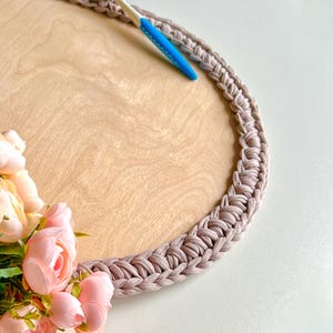 May include: A round wooden tray with a light brown surface and a braided, taupe-colored border. A blue and white crochet hook rests on the tray, near a cluster of pink flowers. The tray is suitable for serving or display.