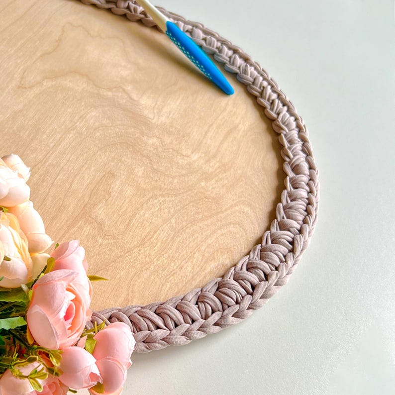 May include: A round wooden tray with a light brown surface and a braided, taupe-colored border. A blue and white crochet hook rests on the tray, near a cluster of pink flowers. The tray is suitable for serving or display.