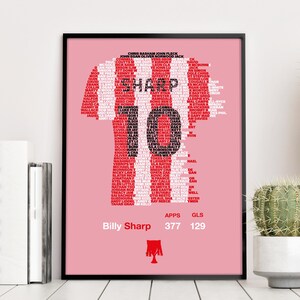 May include: Framed pink print with a red and white striped football shirt design. The shirt is filled with player names, with "SHARP" in the center. Below, it reads "Billy Sharp" with stats: 377 apps, 129 goals. A club crest is at the bottom.