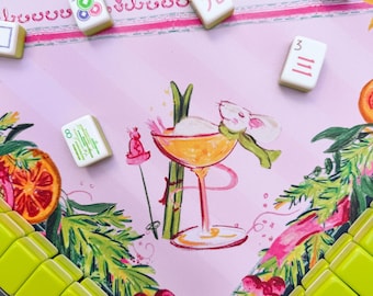May include: A pink mahjong mat featuring a central illustration of a white mouse in a cocktail glass with skis, surrounded by orange slices and pine branches. Several white mahjong tiles with colorful symbols and letters are scattered across the mat.