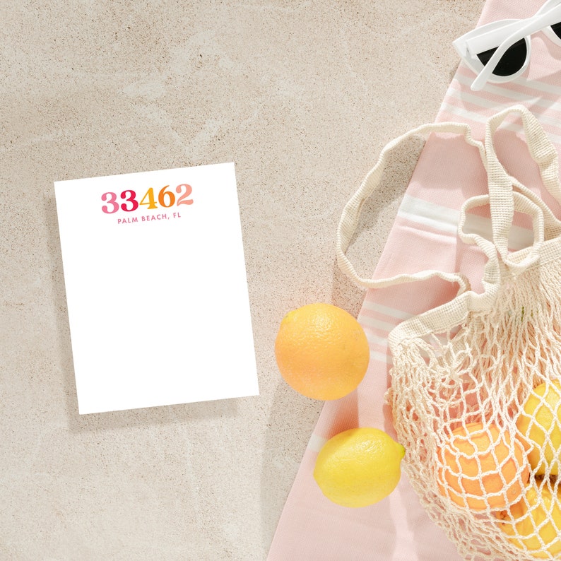 May include: A white notepad with the text "33462 Palm Beach, FL" in pink and orange lettering. The notepad is on a beige surface next to a pink and white striped towel with a white mesh bag filled with oranges and lemons.