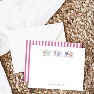 May include: Pink and white striped stationery with a white envelope. The stationery has a pink and white striped border and features four watercolor illustrations of Chinese dominoes with floral designs. The stationery is personalized with the name "Lindsey Jensen".