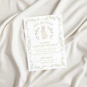 May include: A white bunny with pink ears sits in a green vine border. The text "HOP ON OVER" is above the bunny and "to celebrate LARK'S 1ST BIRTHDAY" is below the bunny. The invitation includes the date, time, location, and contact information for RSVP.