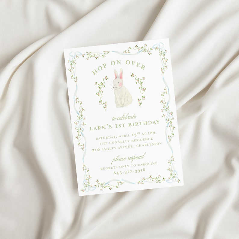 May include: A white bunny with pink ears sits in a green vine border. The text "HOP ON OVER" is above the bunny and "to celebrate LARK'S 1ST BIRTHDAY" is below the bunny. The invitation includes the date, time, location, and contact information for RSVP.