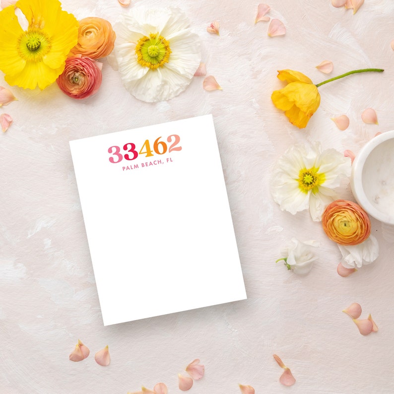 May include: A white notepad with the text "33462 Palm Beach, FL" printed in pink, orange, and yellow. The notepad is surrounded by white and yellow flowers and petals on a pink background.