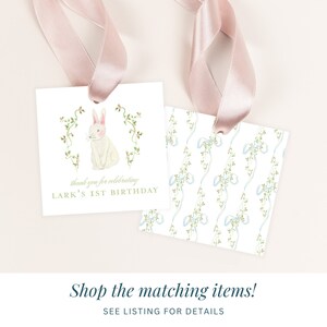 May include: Two white square gift tags with pink ribbon. One tag features a watercolor illustration of a white bunny with pink ears and a green vine border. The text "thank you for celebrating LARK'S 1ST BIRTHDAY" is printed in gold. The other tag features a repeating pattern of blue bows and green vines.