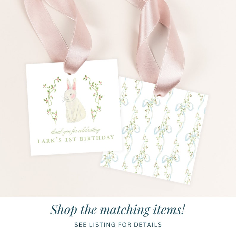 May include: Two white square gift tags with pink ribbon. One tag features a watercolor illustration of a white bunny with pink ears and a green vine border. The text "thank you for celebrating LARK'S 1ST BIRTHDAY" is printed in gold. The other tag features a repeating pattern of blue bows and green vines.