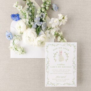 May include: A white envelope with a floral design, containing a white invitation with a green border and a bunny illustration. The invitation reads "Hop on Over to celebrate Lark's 1st Birthday" with details for the event, including the date, time, location, and contact information.