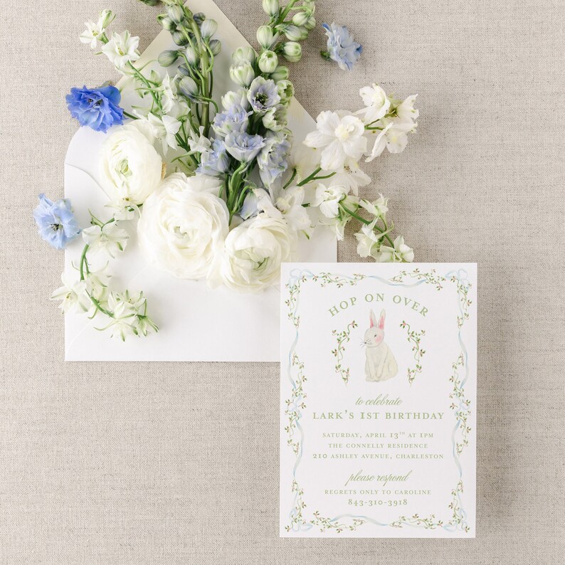 May include: A white envelope with a floral design, containing a white invitation with a green border and a bunny illustration. The invitation reads "Hop on Over to celebrate Lark's 1st Birthday" with details for the event, including the date, time, location, and contact information.