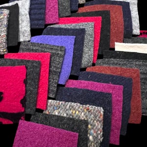May include: A selection of fabric swatches in diverse colours and textures. The swatches feature shades of pink, red, grey, blue, and black. Some swatches have a textured appearance, while others are smooth. The arrangement is layered, displaying the variety of materials.