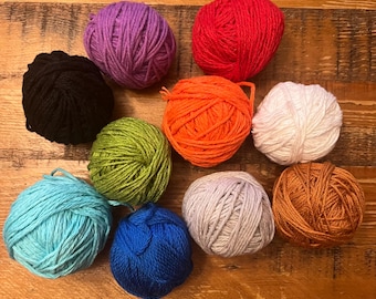 May include: A collection of colorful yarn balls in various shades, including purple, red, orange, white, brown, black, green, blue, and light blue. The yarn balls are arranged on a wooden surface, ideal for crafting projects.