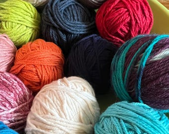 May include: A collection of colorful yarn balls in various shades, including green, blue, red, orange, and white. The yarn is tightly wound, suggesting it's suitable for knitting or crochet projects. The balls are arranged in a green container.