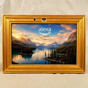 Op de afbeelding: Een rechthoekig, goudkleurig omlijst scherm met een schilderachtig landschap met bergen, een meer en bomen. Het woord "alexa" wordt in het midden weergegeven, met het Amazon-logo erboven. De lijst heeft een gestructureerd, sierlijk ontwerp.