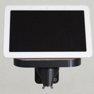 Könnte beinhalten: Ein weißes Smart Display mit einem schwarzen Bildschirm, das an einer schwarzen Wandhalterung montiert ist. Die Halterung hat eine kleine Ablage, auf der das Display stehen kann.