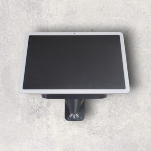 Peut inclure: Une tablette rectangulaire avec un écran noir et un cadre blanc est fixée au mur. La tablette est fixée à un support noir avec une base pivotante. Le mur est de couleur gris clair.