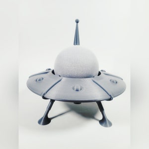 May include: A gray, 3D printed UFO-shaped speaker with a round, gray speaker on top. The speaker has three legs.