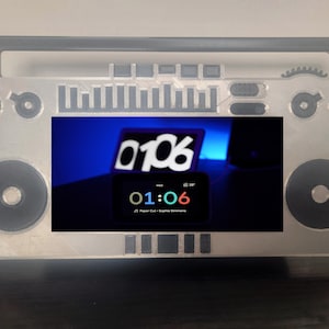 Op de afbeelding: Een zilverkleurig frame in de vorm van een boombox met een digitaal display met de tijd 01:06. Het display is zwart met witte, gele en rode tekst. De boombox heeft een handvat, luidsprekerdetails en bedieningsknoppen.