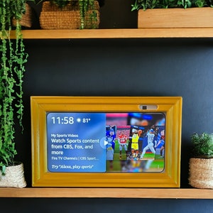Könnte beinhalten: Ein gelber digitaler Bilderrahmen mit einem Bildschirm, der die Zeit 11:58 und die Temperatur 81 Grad anzeigt. Der Bildschirm zeigt auch ein Sportvideo mit dem Text "My Sports Videos Watch Sports content from CBS, Fox, and more Fire TV Channels | CBS Sport... Try "Alexa, play sports""