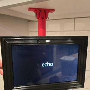 Op de afbeelding: Een zwart omrand apparaat met het woord "echo" op het scherm, gemonteerd op een rode steunstructuur. Het apparaat is bevestigd aan de onderkant van een witte plank.