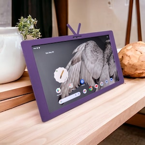 Puede incluir: Una tableta de color morado con una pantalla que muestra un ala de pájaro gris y la fecha "Sáb, 20 de mayo". La tableta está sobre una superficie de madera.