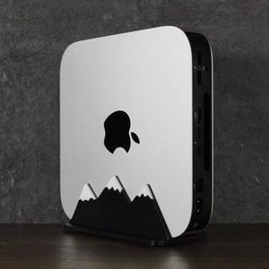 Op de afbeelding: Een witte Apple Mac mini computer met een zwart Apple-logo aan de voorkant. De computer staat op een zwarte standaard met een bergontwerp.