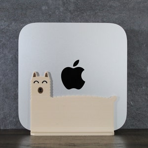 May include: A white computer with an Apple logo sits on a beige llama-shaped stand. The stand has a brown nose and black eyes.