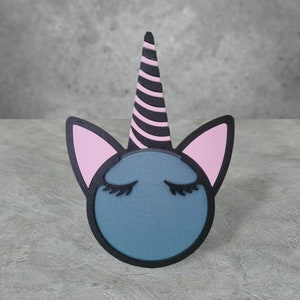 Puede incluir: Una cabeza de unicornio azul con orejas rosas y un cuerno rayado en blanco y negro. El unicornio tiene los ojos cerrados con pestañas negras.