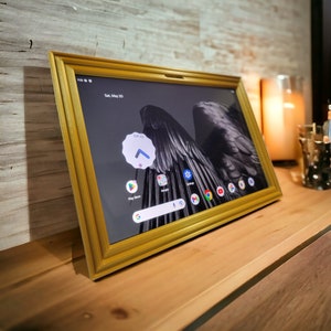 Puede incluir: Un marco de fotos digital dorado con una imagen en blanco y negro de alas en la pantalla. La pantalla muestra la fecha "Sáb, 20 de mayo" y varios iconos de aplicaciones, incluidos Google Play Store, Google y Gmail.