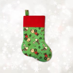 May include: A Christmas stocking with a green body featuring a pattern of Charlie Brown and Snoopy characters. The top of the stocking is red, and a green loop is attached for hanging. Candy canes and wreaths are also part of the pattern.