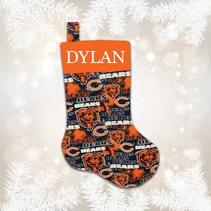 May include: A personalized Christmas stocking with the name "DYLAN" embroidered on the cuff. The stocking is decorated with a repeating pattern of the Chicago Bears logo, featuring a bear head and the letter "C" in orange and black.