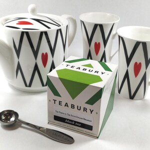 May include: A white ceramic tea set with a teapot, two mugs, and a box of tea. The teapot and mugs feature a black and white geometric pattern with red heart accents. A silver tea scoop is also included.