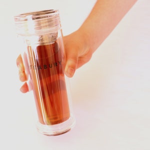 May include: A clear glass tea bottle with a stainless steel lid and infuser. The bottle is filled with brown tea and has the word "TEABURRY" printed on the side.