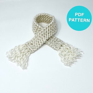 May include: A cream-colored, knitted scarf with fringed ends. The scarf is folded in half, with the ends hanging down. A turquoise circle with the text "PDF PATTERN" is in the upper right corner. The scarf is made of a soft, textured yarn.